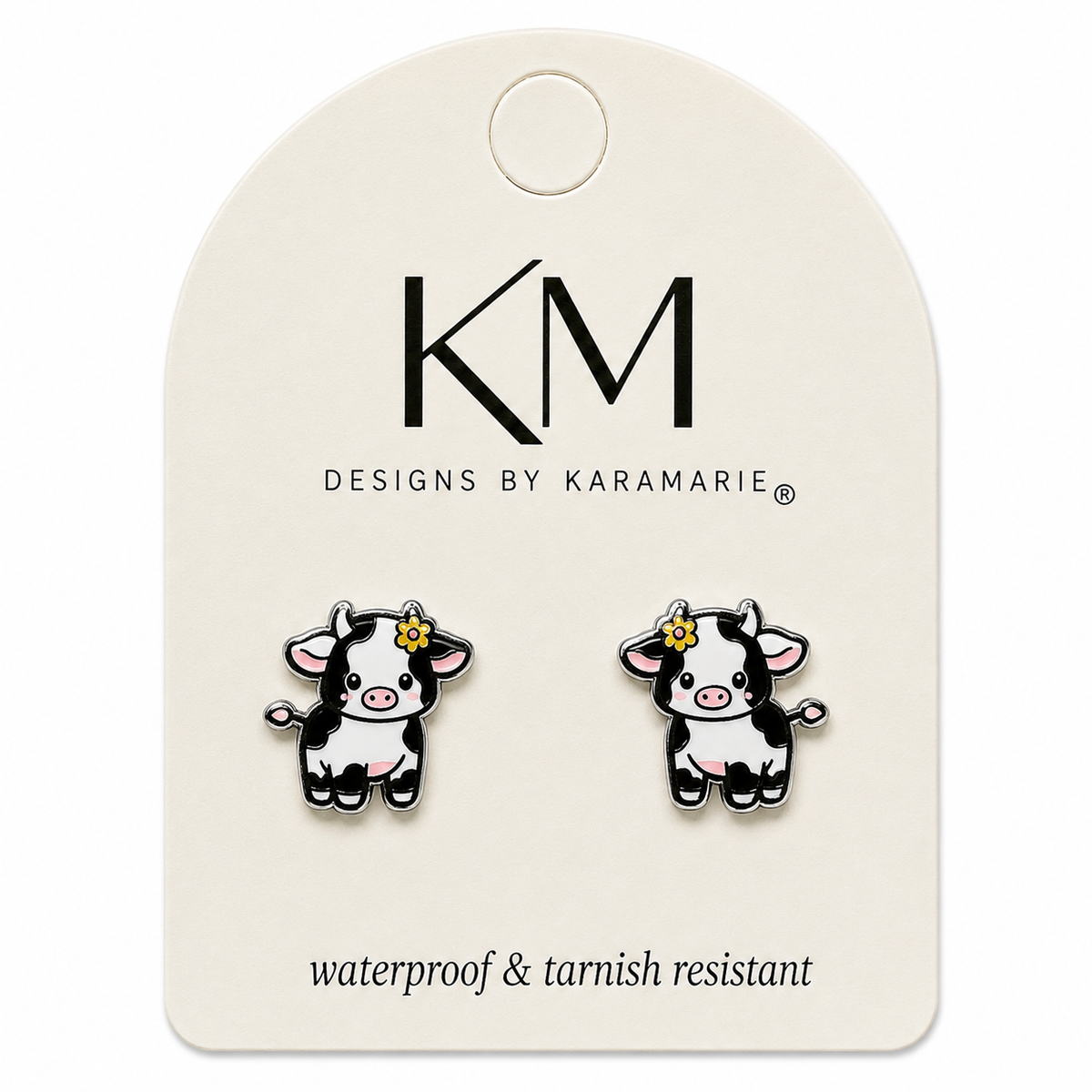 A pair of cow stud earrings displayed on a cream-colored branded caThe card features the logo 'KM DESIGNS BY KARAMARIE