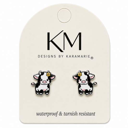 A pair of cow stud earrings displayed on a cream-colored branded caThe card features the logo 'KM DESIGNS BY KARAMARIE